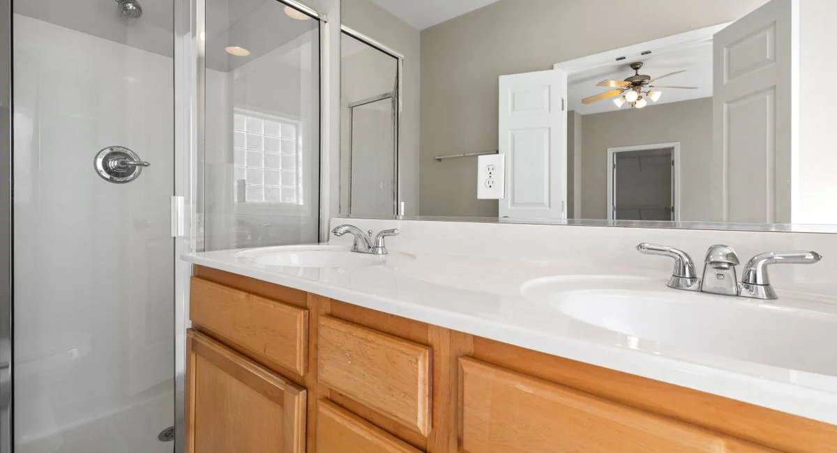 Bright and modern bathroom featuring a dual sink vanity, glass shower, and large mirror reflecting ceiling fan light.