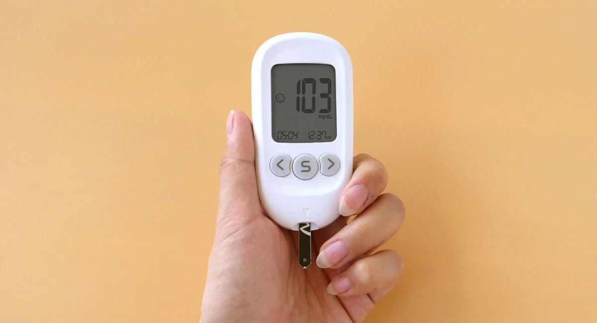A hand holding a blood glucose meter displaying 103 mg/dL on a neutral background.