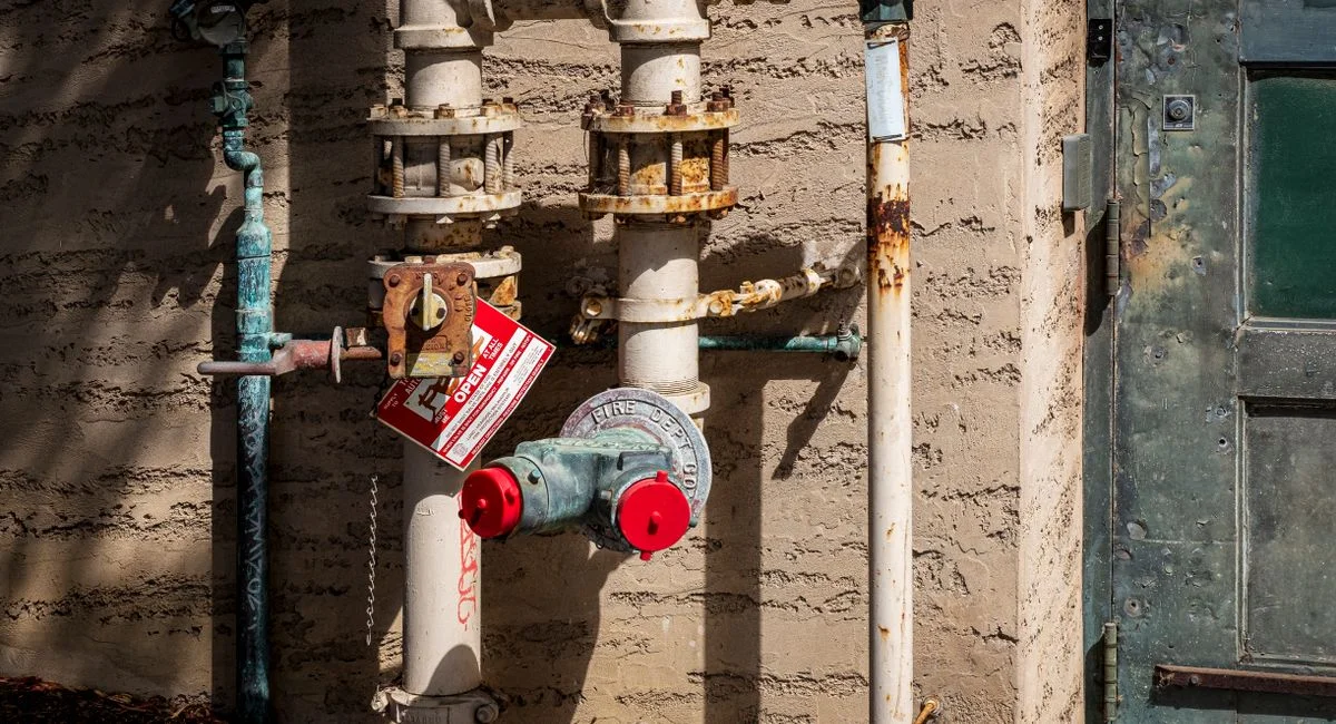 Rusty outdoor plumbing pipes with pressure gauges and warning signs on a wall.