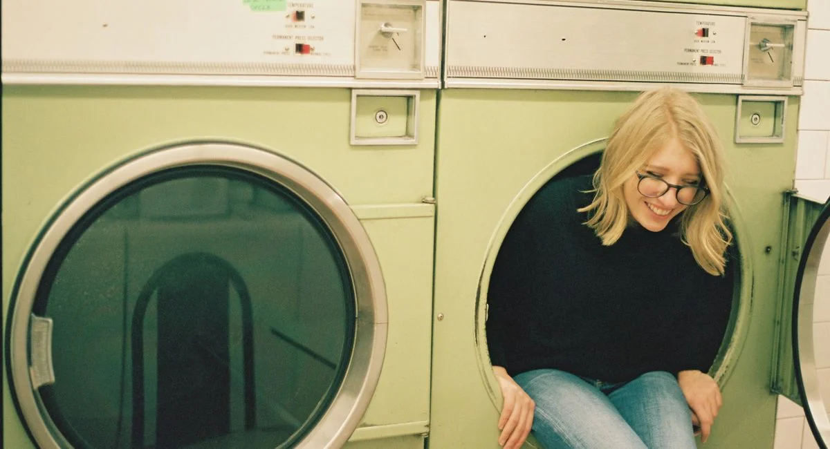 Positive blond female in eyeglasses wearing black sweater sitting in tub of washing machine while having fun in laundry room