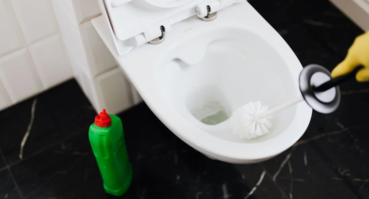 From above of crop anonymous person in rubber gloves using liquid toilet sanitizer and brush to clean toilet bowl in bathroom